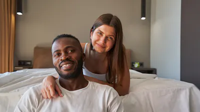 Smiling woman lying on bed and hugging pleased man seated on floor